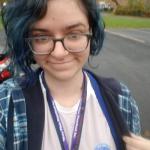 a photo of a woman with dyed blue hair and glasses, wearing a blue plaid jacket and a light purple shirt